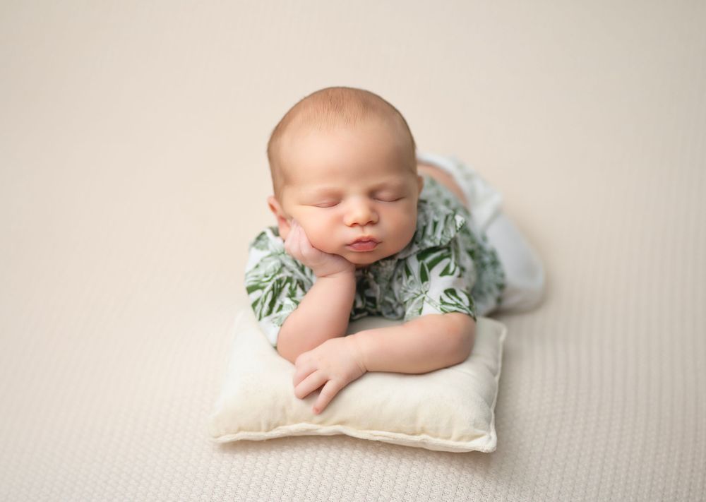 Sleeping newborn in pale floral outfit rests peacefully on small white pillow during professional photo session.
