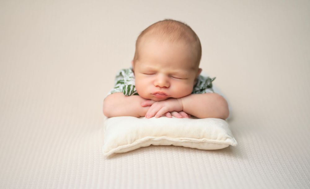 Sleeping newborn rests peacefully with folded arms on white pillow with greenery accent in studio setting.
