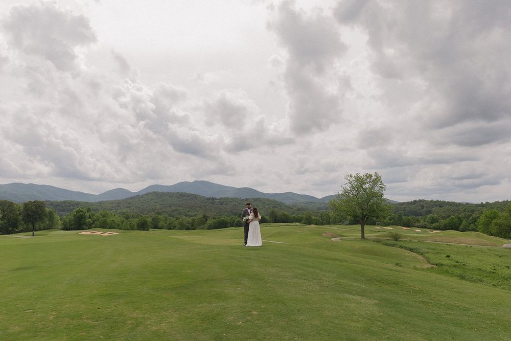 A couple stands on a lush green field in chattanooga with mountains and dramatic cloudy skies in the background.