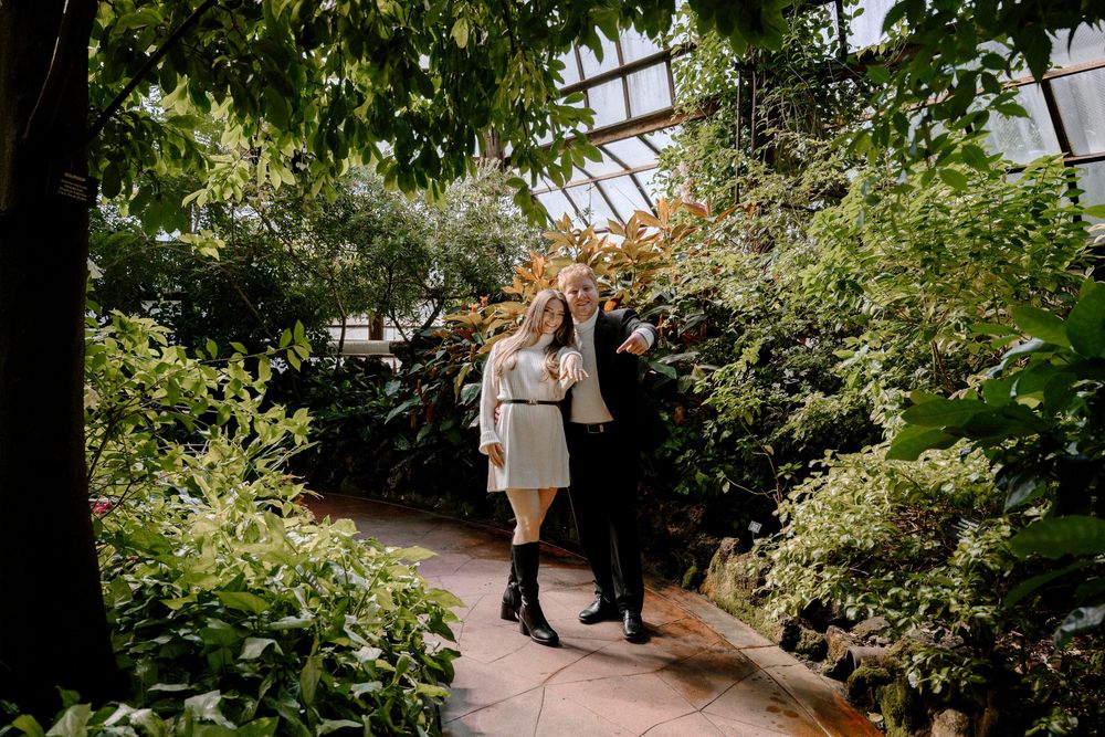 A couple walks together holding hands along a lush greenhouse pathway surrounded by green plants and natural light.