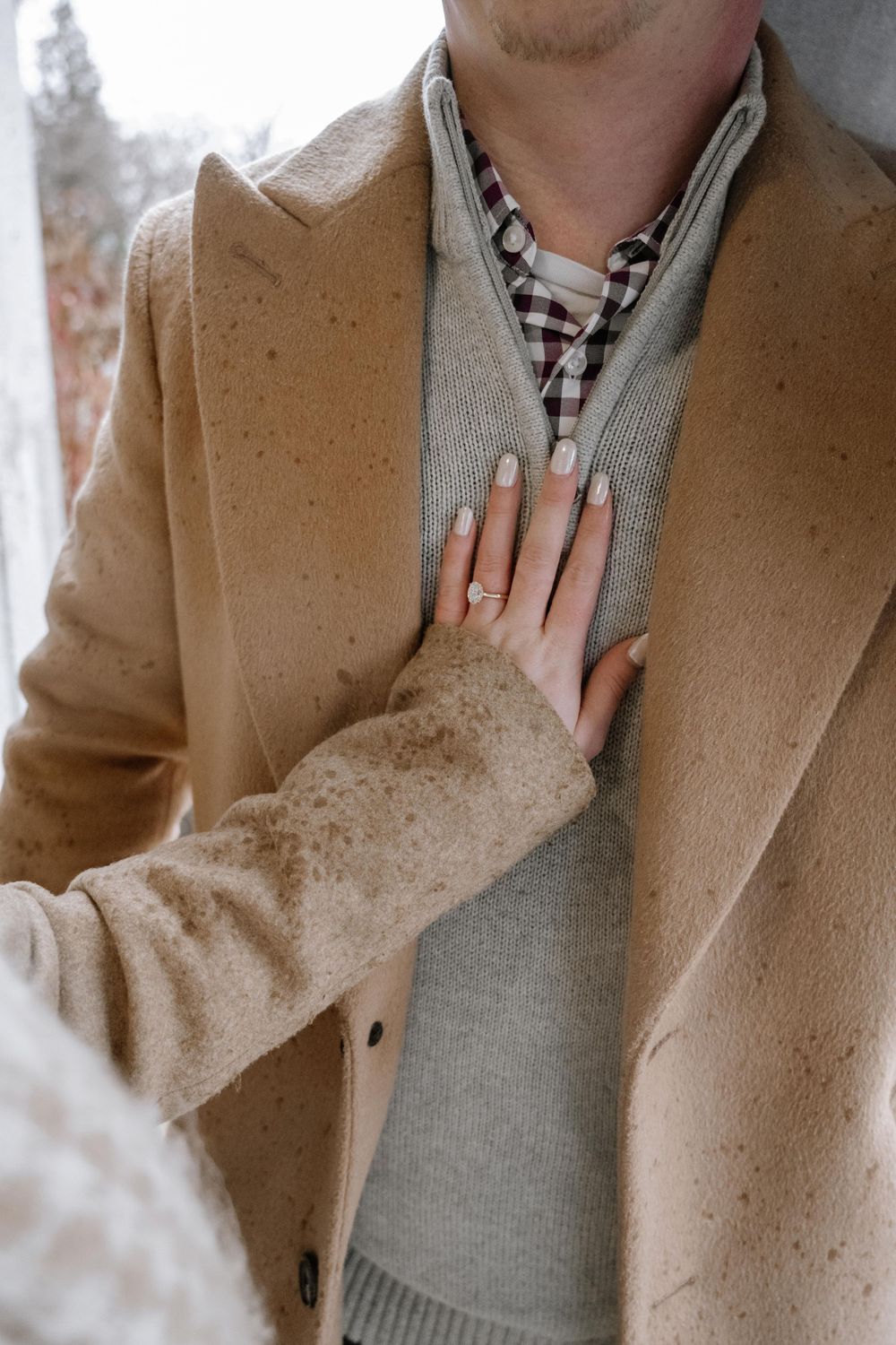 Close-up of someone wearing layers of tan coat, gray sweater and plaid shirt with hand resting on chest.