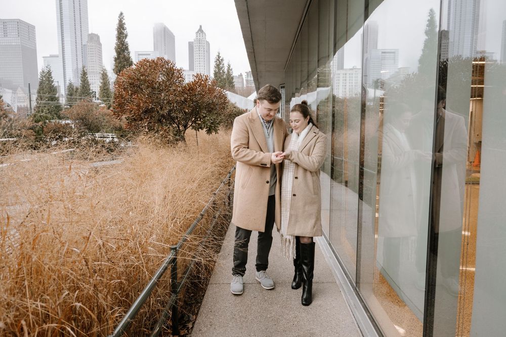 A couple in matching beige coats stands together on an urban path with tall grass and city buildings in the background.