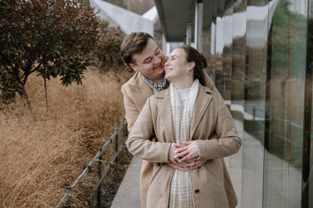 Two people in beige coats embrace lovingly against a modern glass building exterior with autumn grasses.