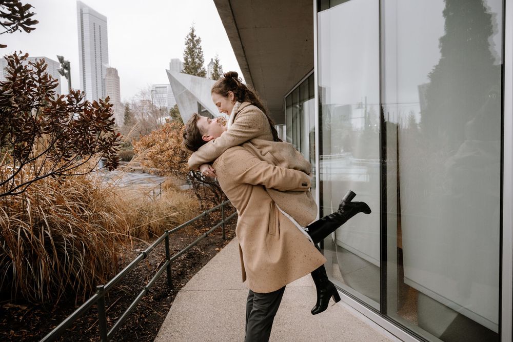 A person wearing a beige coat holds a small dog while standing on a balcony with urban scenery in the background.