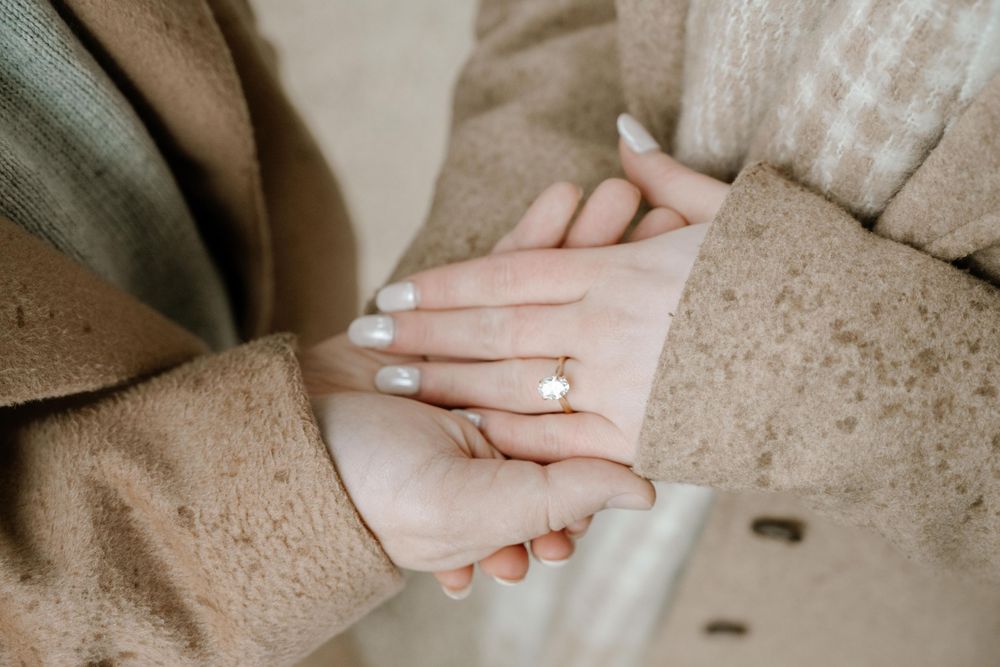 Two hands clasped together in an elegant engagement or wedding pose on a beige textured background.