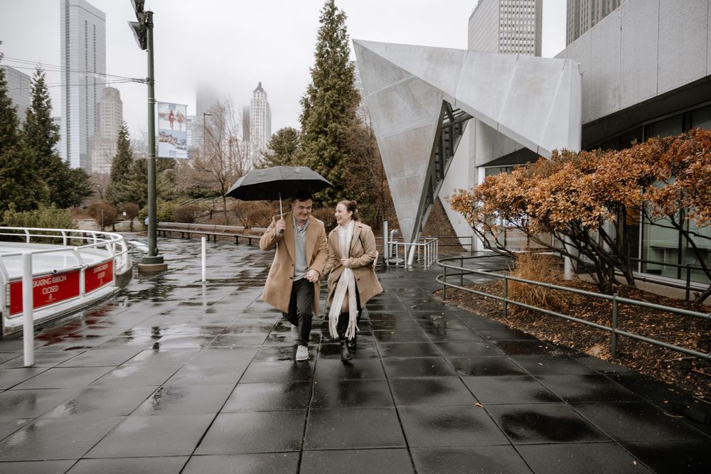 Two people share an umbrella while walking on wet pavement near modern architecture buildings on a rainy autumn day in downtown.