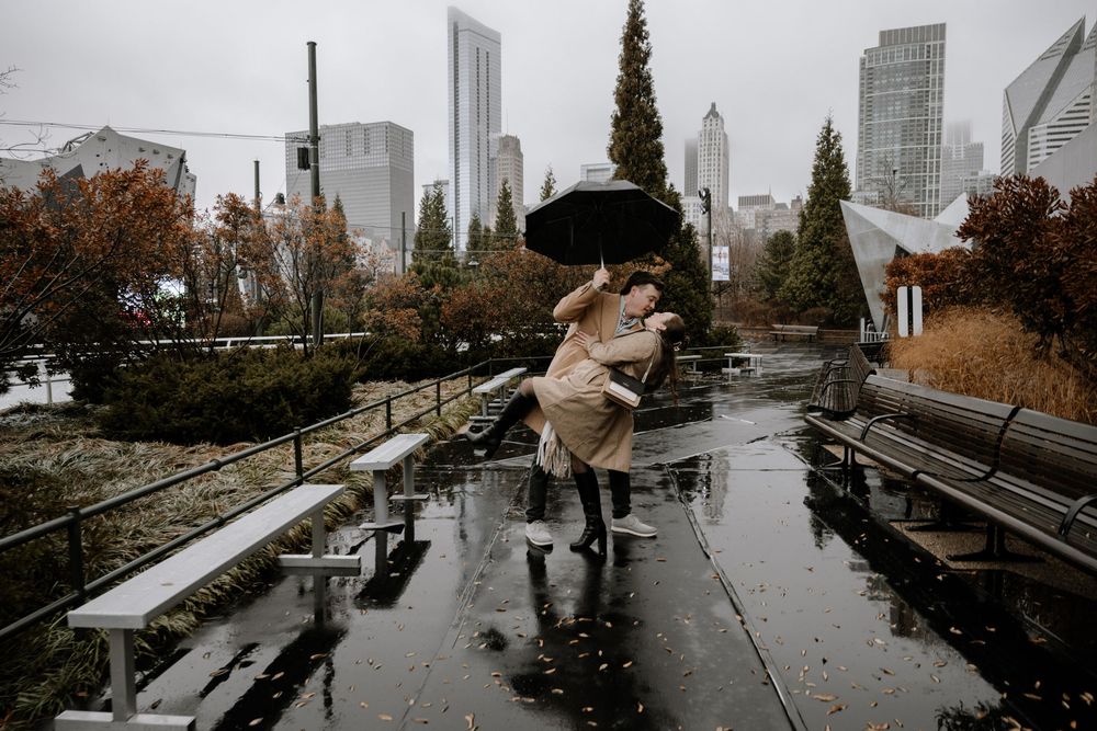 Someone leaps over a puddle with an umbrella in a rainy urban park with modern skyscrapers in the background.