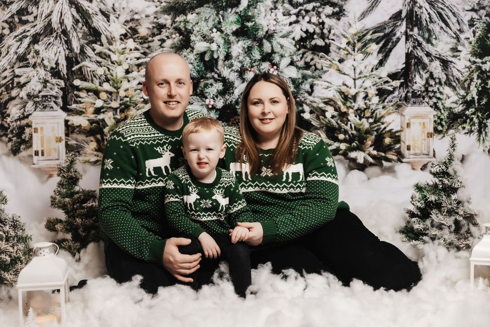 A family wearing matching green holiday sweaters poses together against a snowy winter backdrop with evergreen trees.