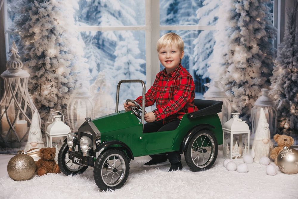 Child in red plaid shirt rides green vintage pedal car in winter wonderland setting with snow and decorated trees.