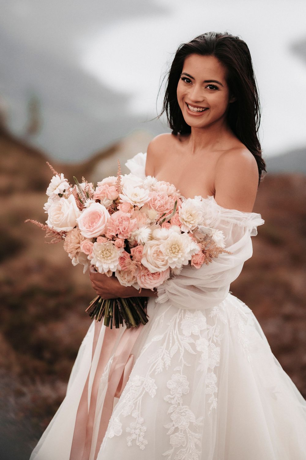 A bride in an off-shoulder wedding gown holds a romantic blush pink and white floral bouquet against a moody landscape.