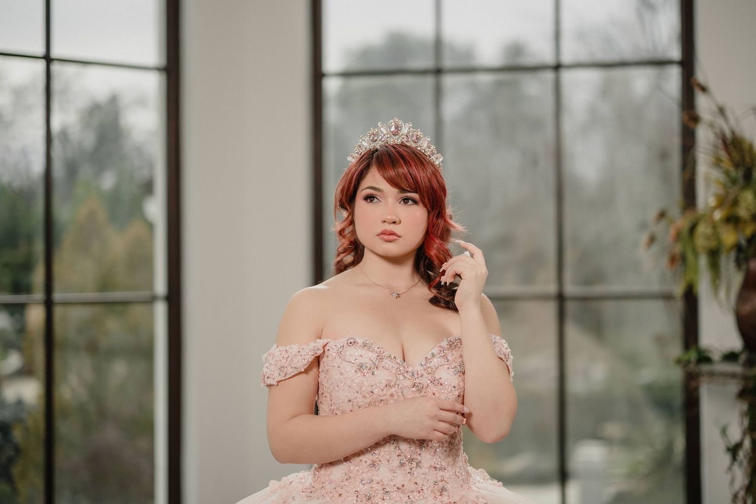 A quinceanera in a soft pink off-shoulder gown poses elegantly in front of large windows with natural light.