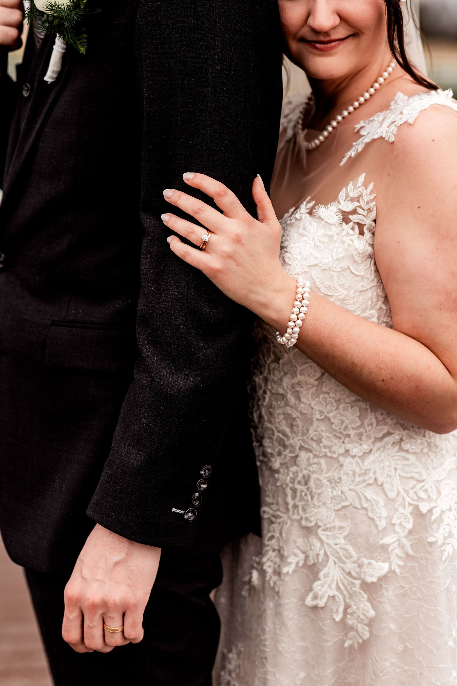 Close-up wedding photo of bride holding groom’s arm, showing lace dress, pearl jewelry, and wedding rings.
