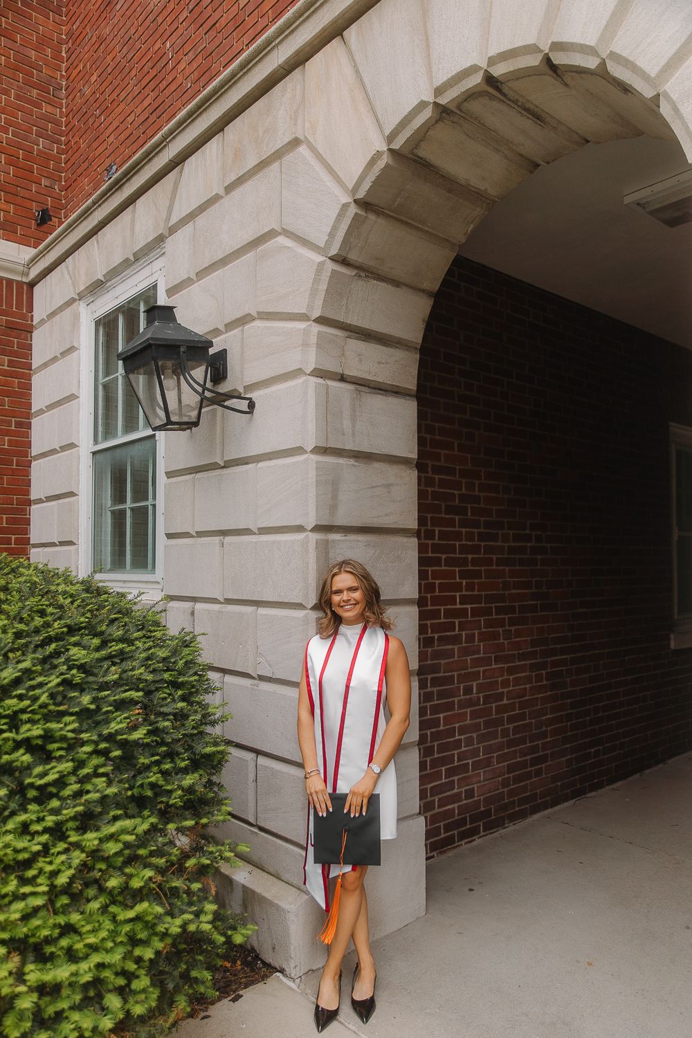Graduate wearing a white halter dress and black cap makes a peace sign on a sunny path with brick buildings behind.