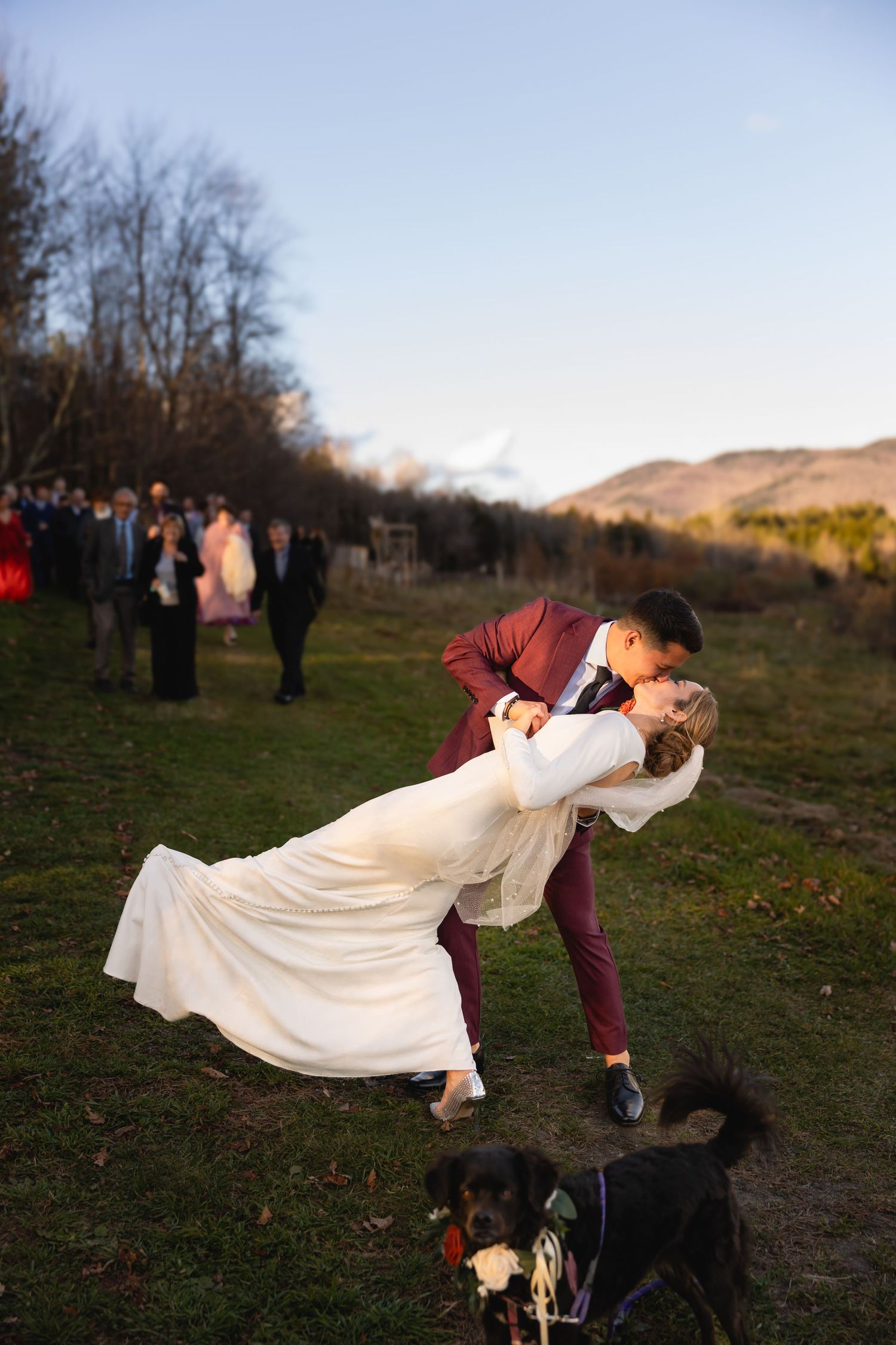 Couple shares a romantic dip pose during sunset wedding celebration while their dog watches nearby in mountain landscape.