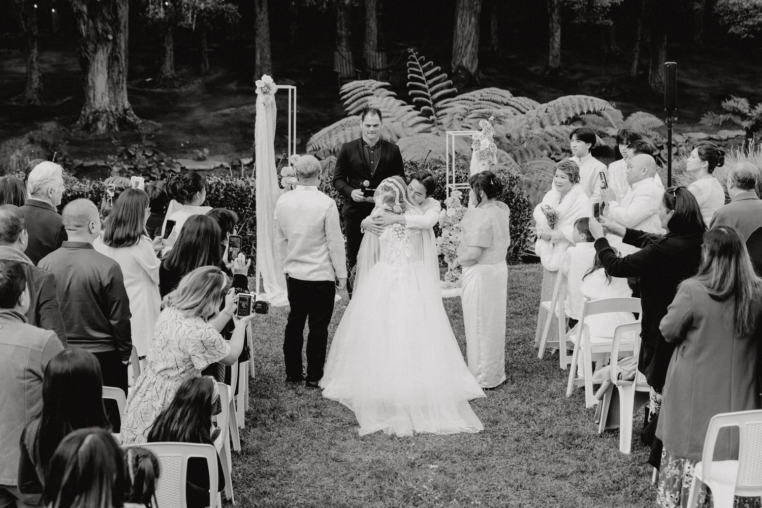 A black and white photograph of a garden wedding ceremony with guests seated on white chairs.