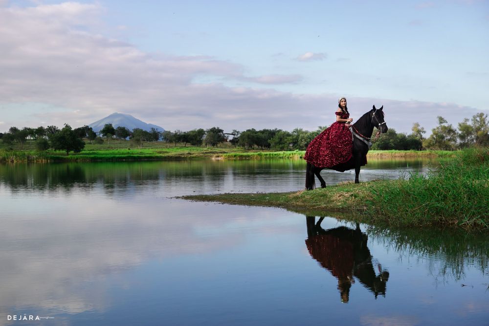 A black horse with rider in red dress stands beside a reflective lake with mountains in the background.
