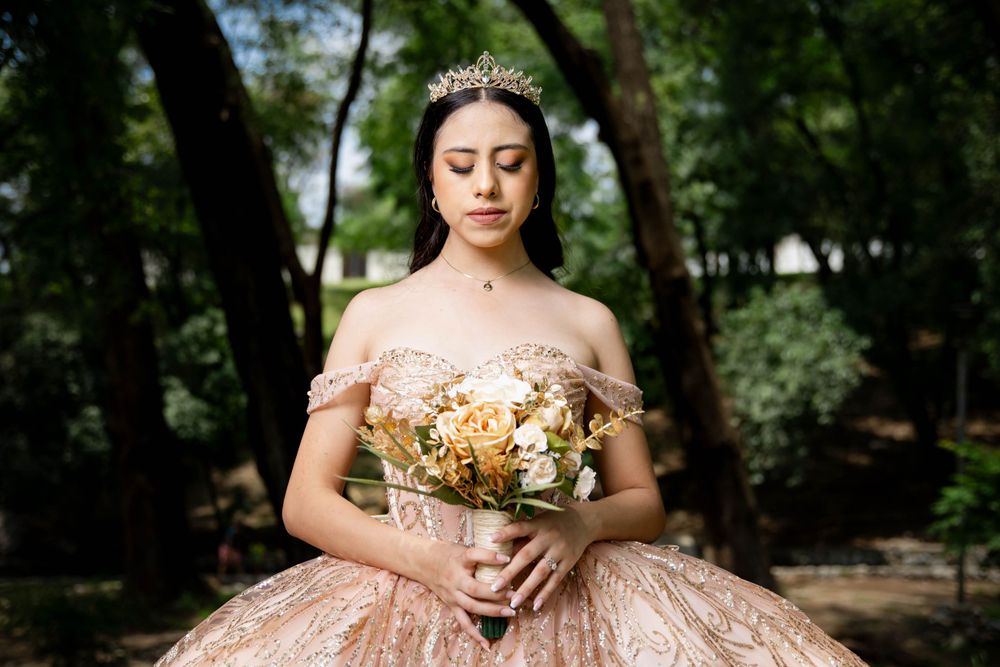 A sparkling pink quinceañera gown glimmers in a wooded setting while holding a bouquet of dried flowers.