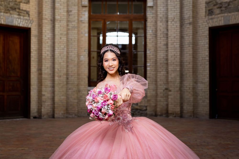 Elegant pink quinceañera dress with floral bouquet photographed in front of ornate stone church entrance.