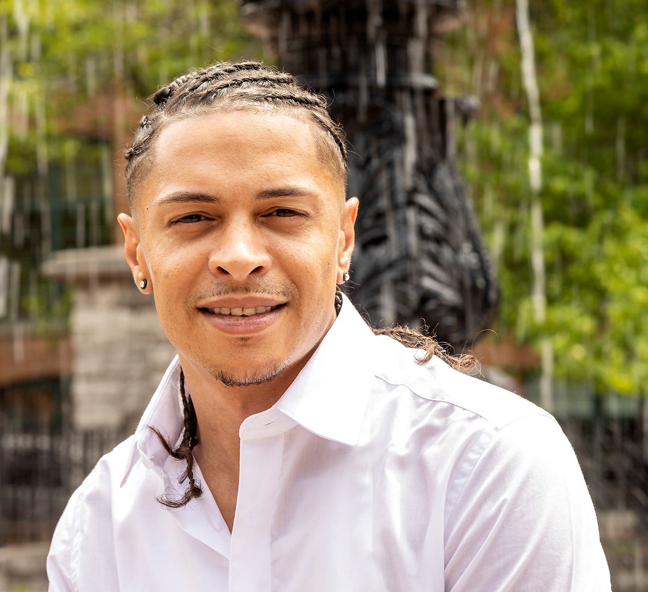 Young man in white dress shirt smiles warmly while posing outdoors with greenery in background.