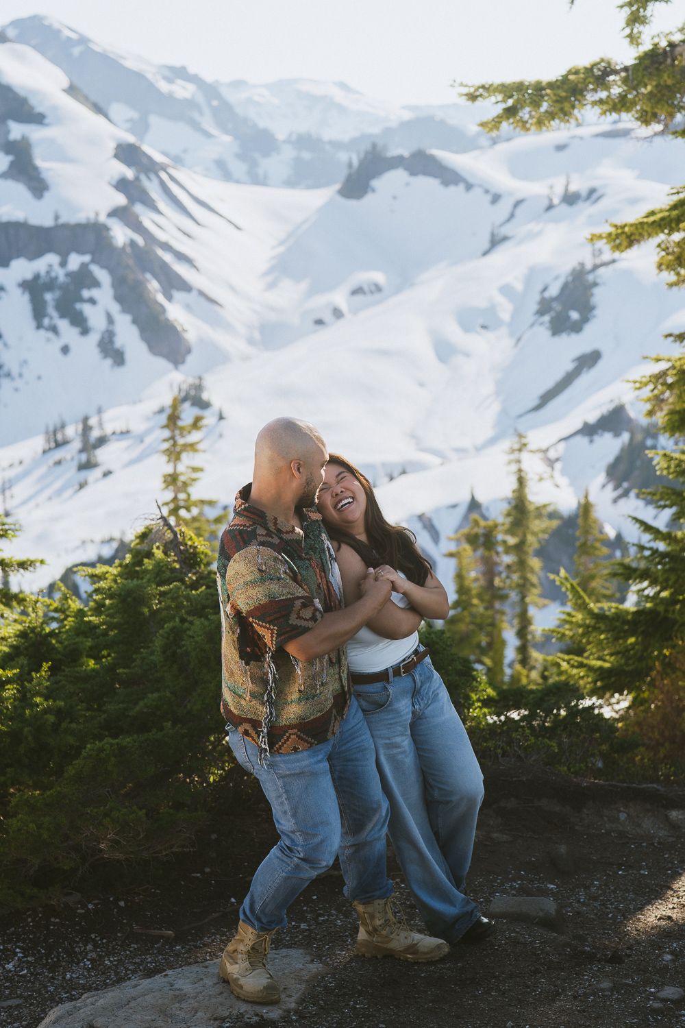 Two people in denim share an intimate moment against a snow-capped mountain backdrop with evergreen trees.