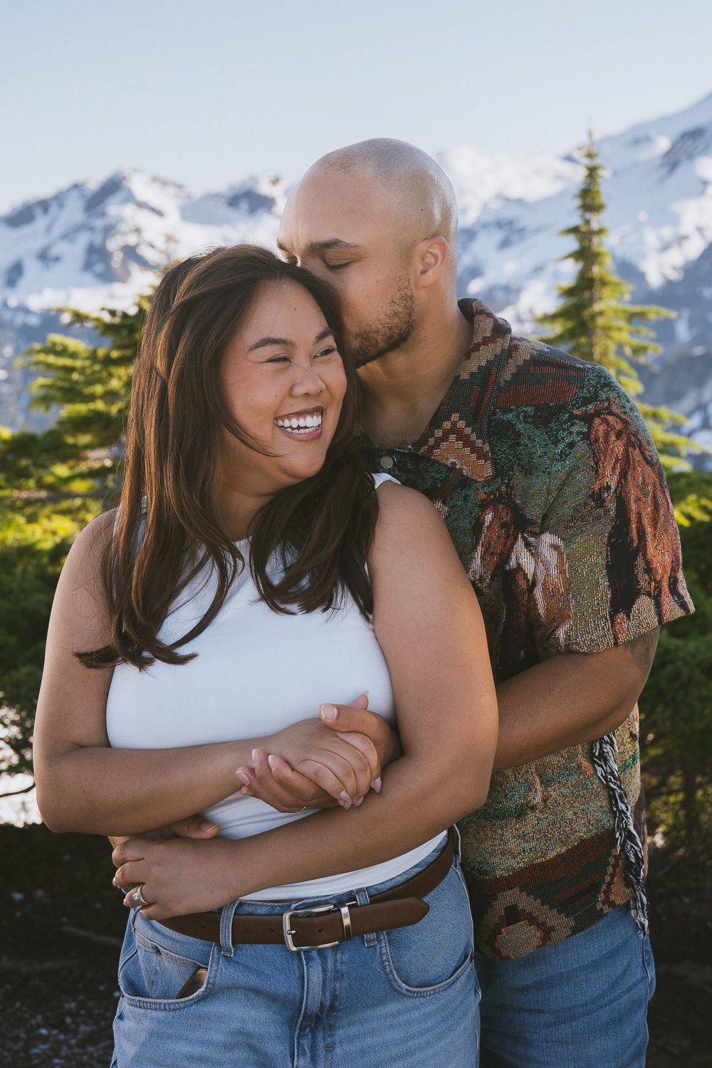 Joyful couple embracing and laughing together in front of snowy mountain peaks and evergreen trees.