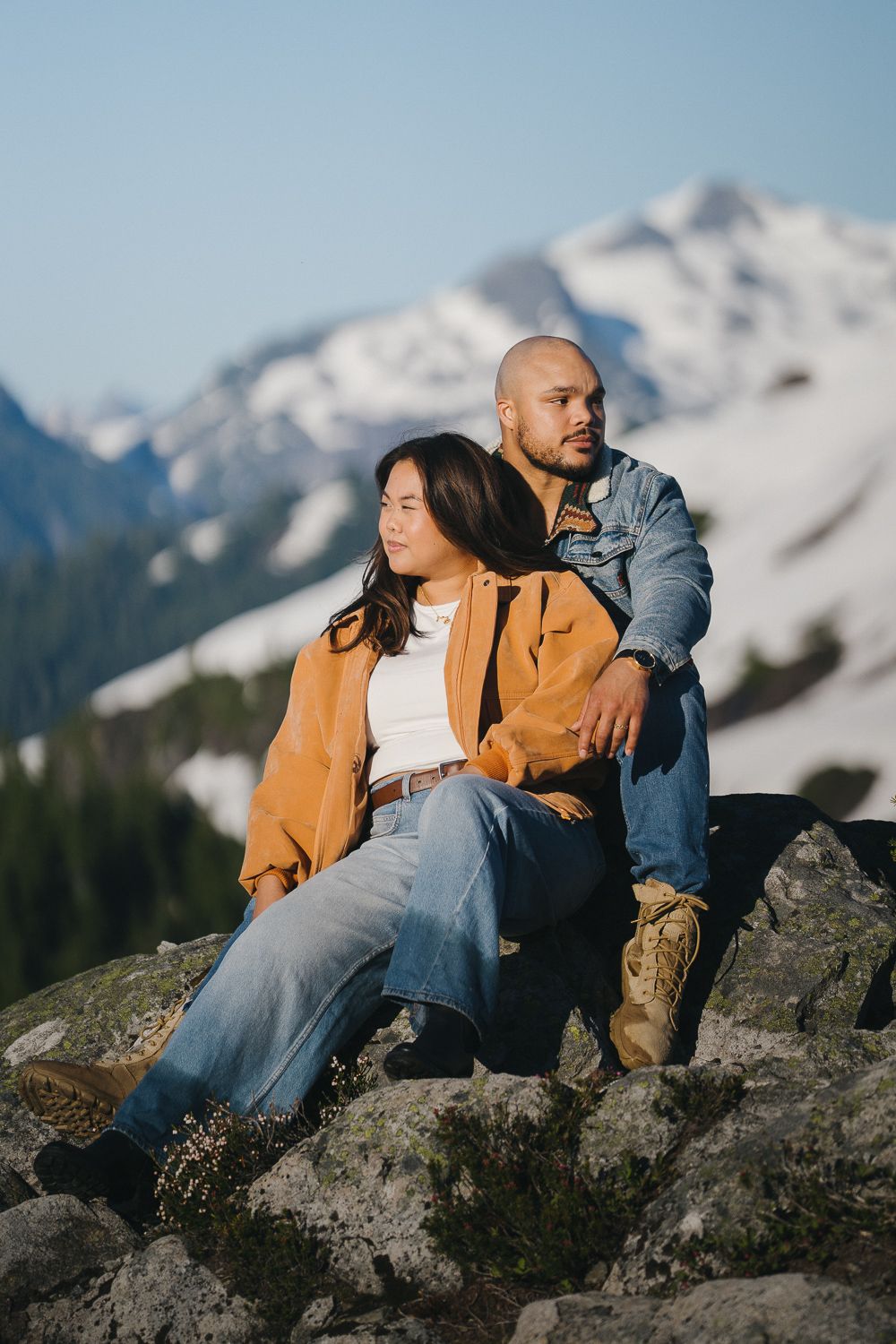 A couple sits together on rocky outcrops with snow-capped mountain peaks visible in the background on a clear day.