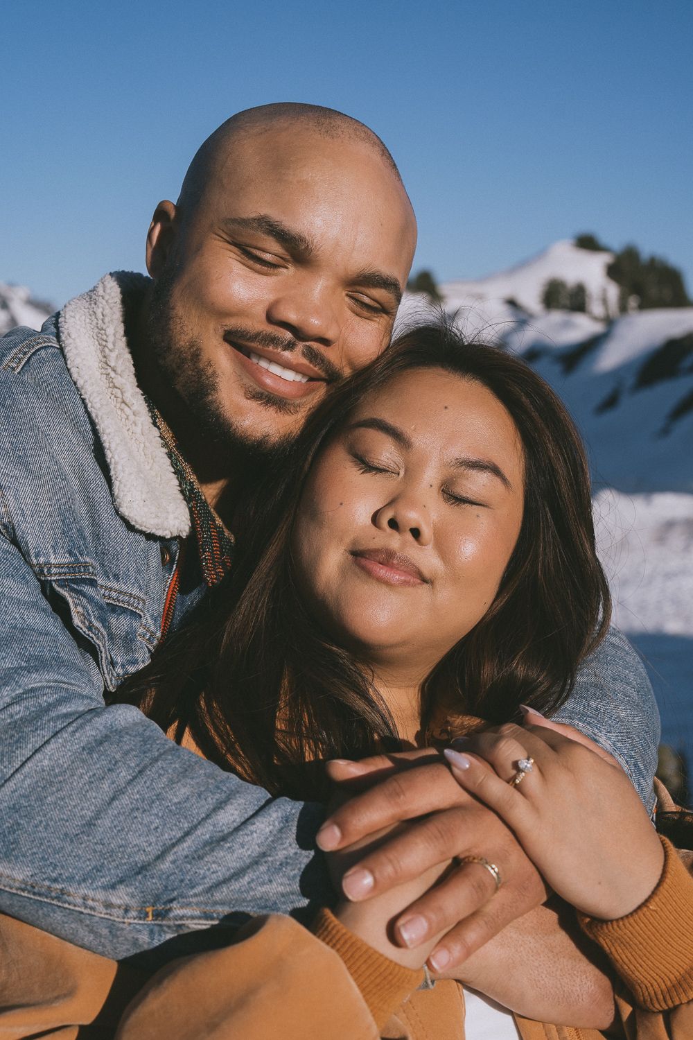 A loving couple embraces while posing playfully in front of a snowy mountain backdrop on a sunny winter day.