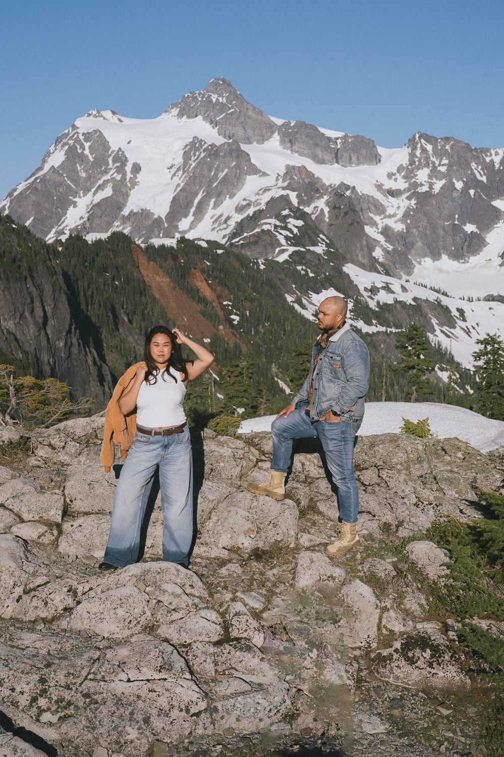 Two travelers enjoy a scenic overlook on rocky terrain with snow-capped mountains in the background.