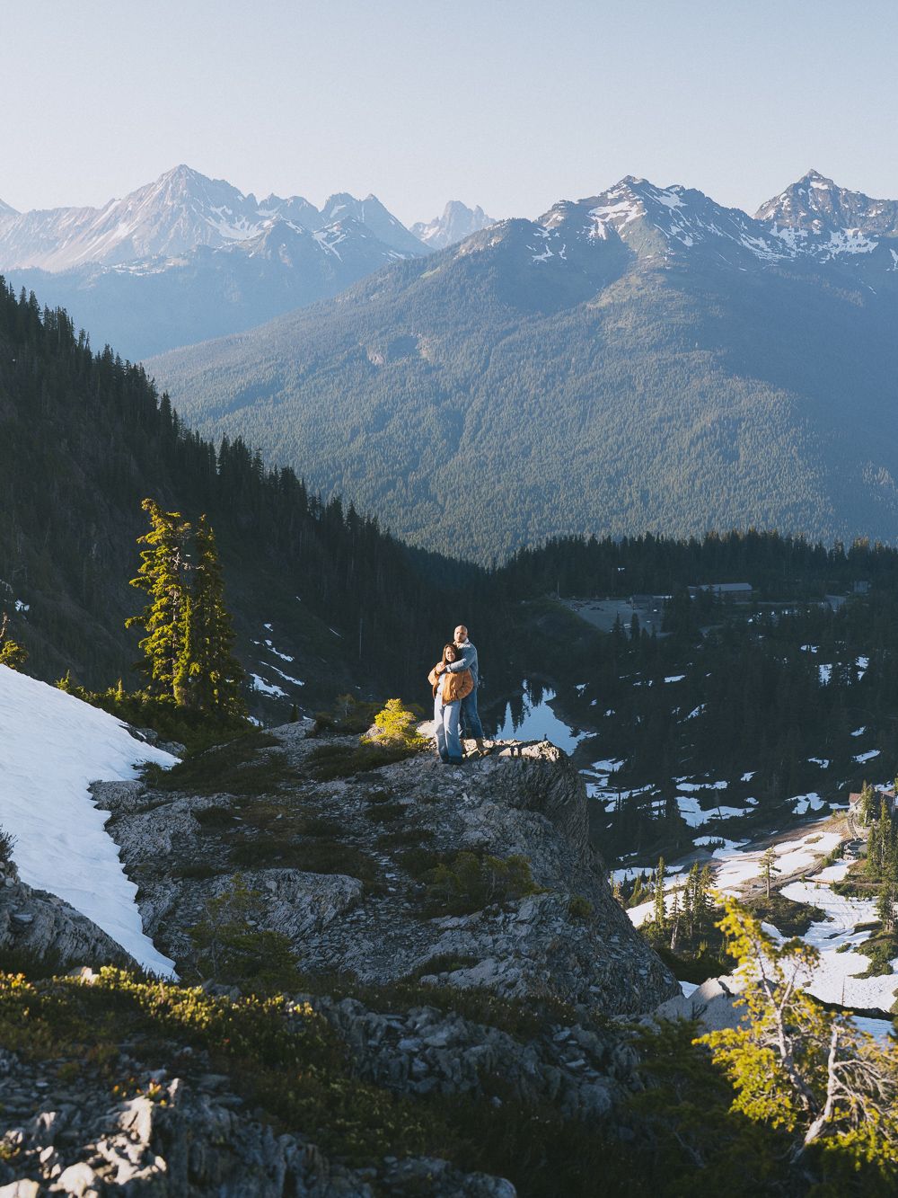 A figure stands on a snowy mountain ledge overlooking a scenic mountain range with evergreen forests in Washington state.