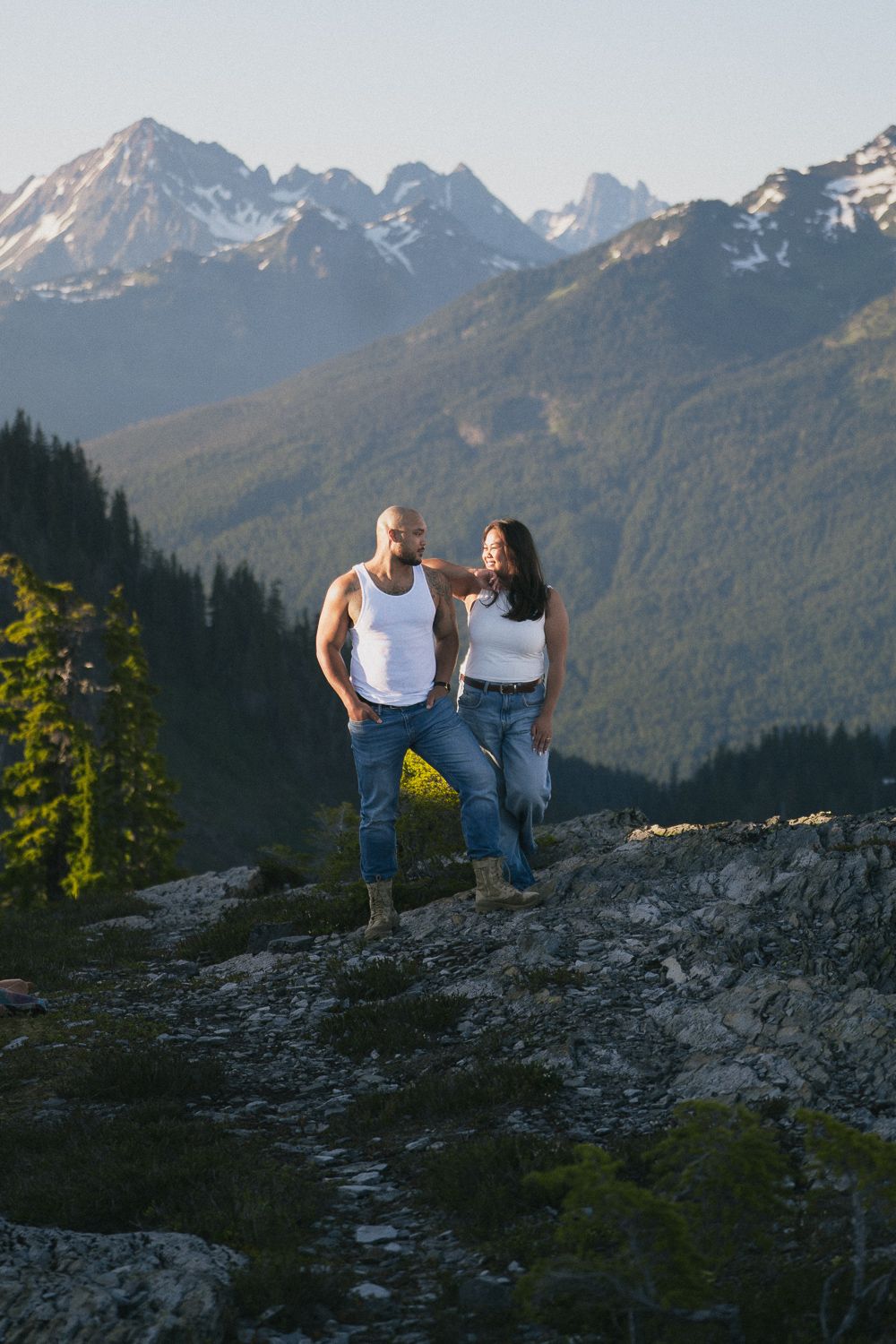 A couple stands together on a rocky mountain overlook with snow-capped peaks and evergreen forests in the background.