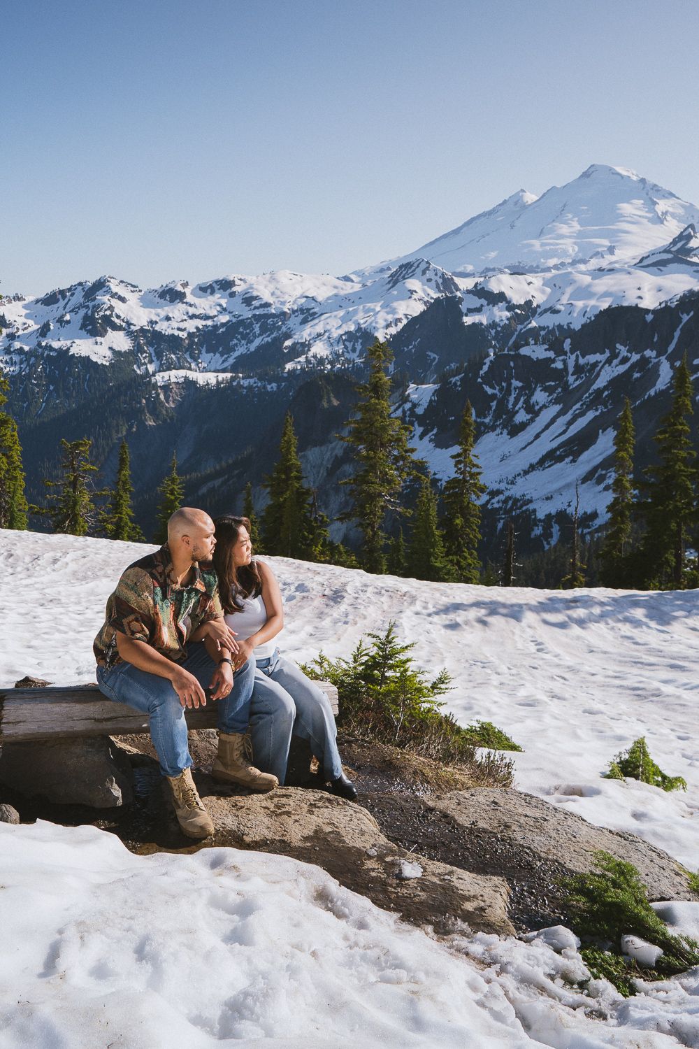 Couple sits on rocky outcrop overlooking snowy Mount Baker wilderness with evergreen trees and mountain peaks in Washington.