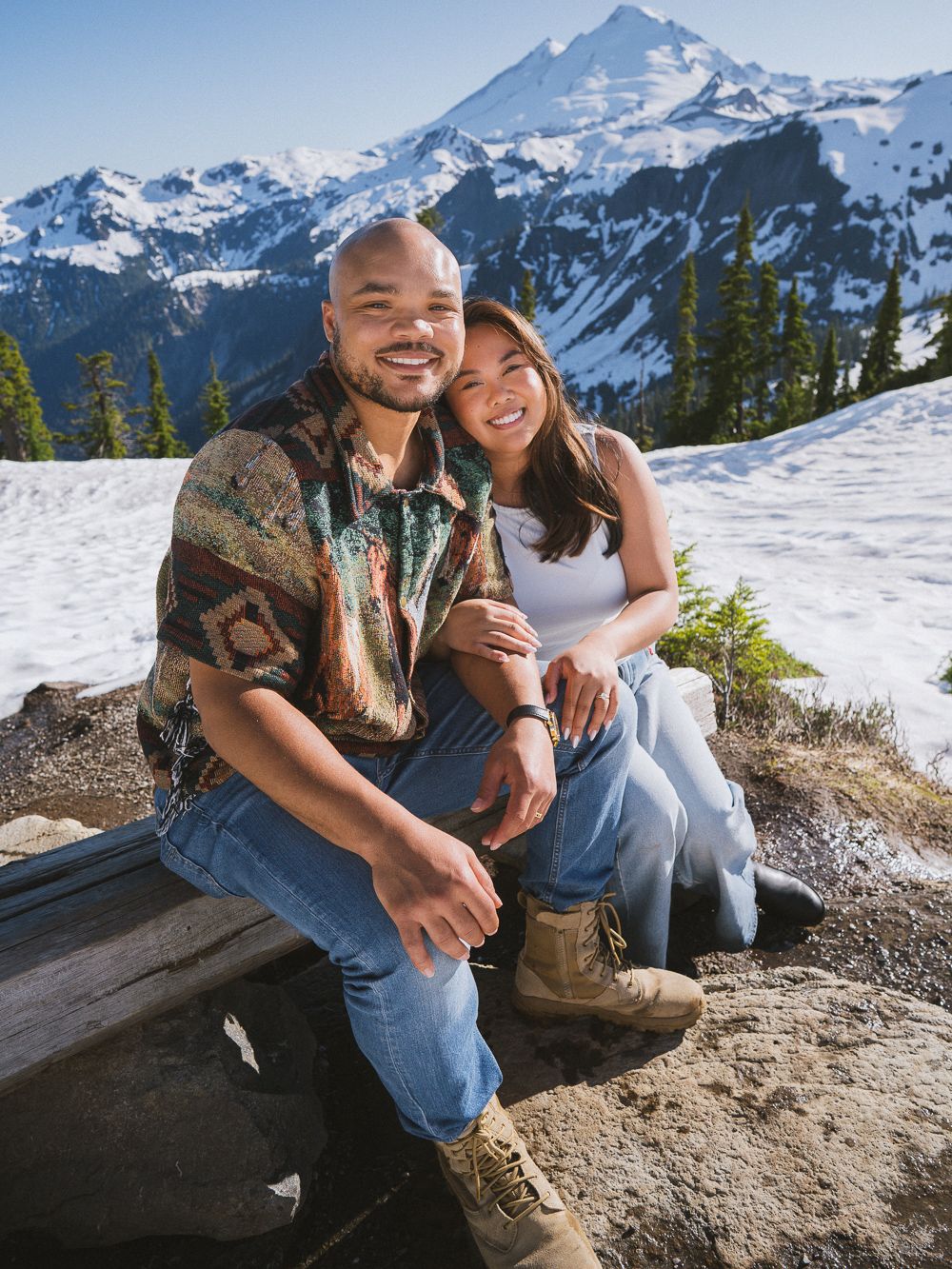 Snow-capped mountain peaks tower over a couple sitting together on a rocky outcrop during a winter adventure.