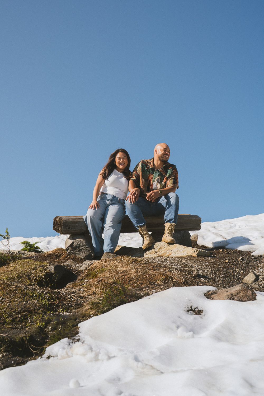 Couple resting on snowy mountain edge with their dog enjoying a bright winter day against blue sky.