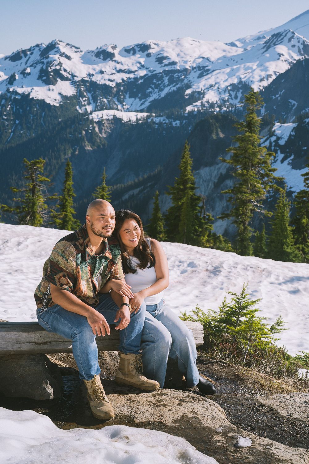 A couple sits together on a rocky ledge overlooking snow-capped mountains and evergreen forest in winter.
