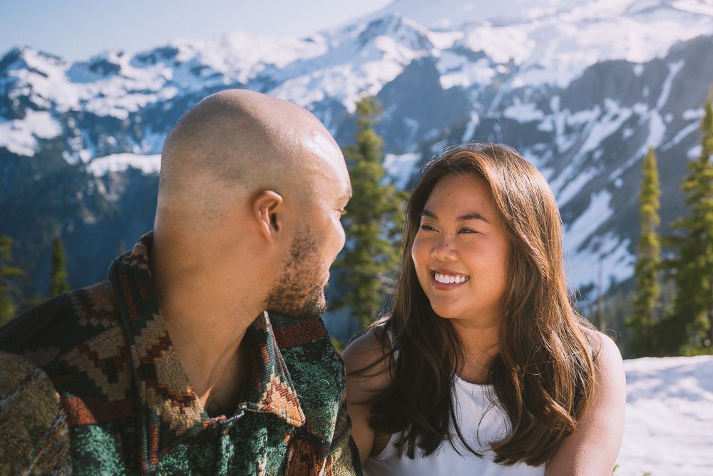 A couple shares a joyful moment while sitting in the snow with majestic snow-capped mountain peaks in the background.