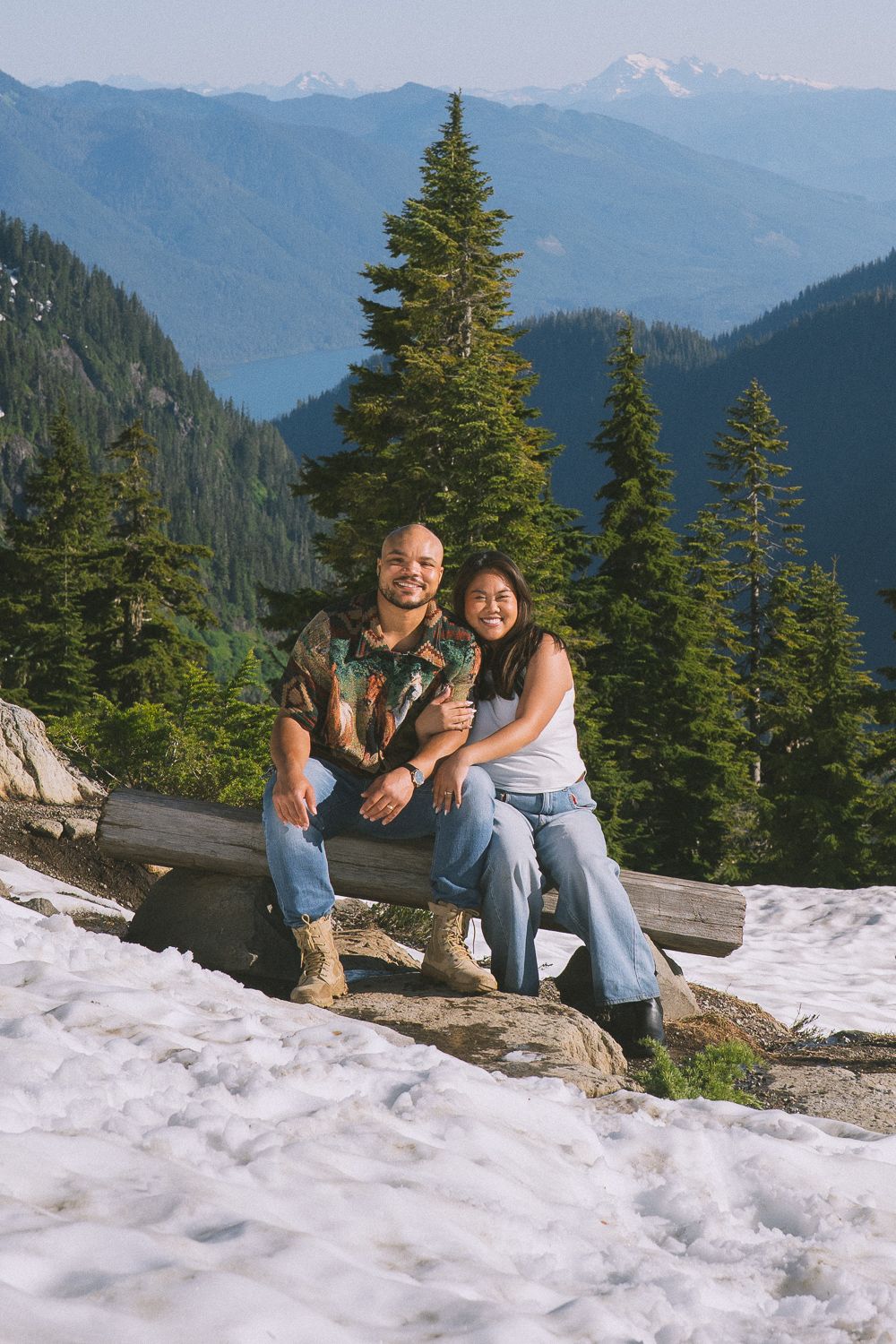 A couple sits together on a rocky outcrop surrounded by snow and evergreen trees in a scenic mountain landscape.
