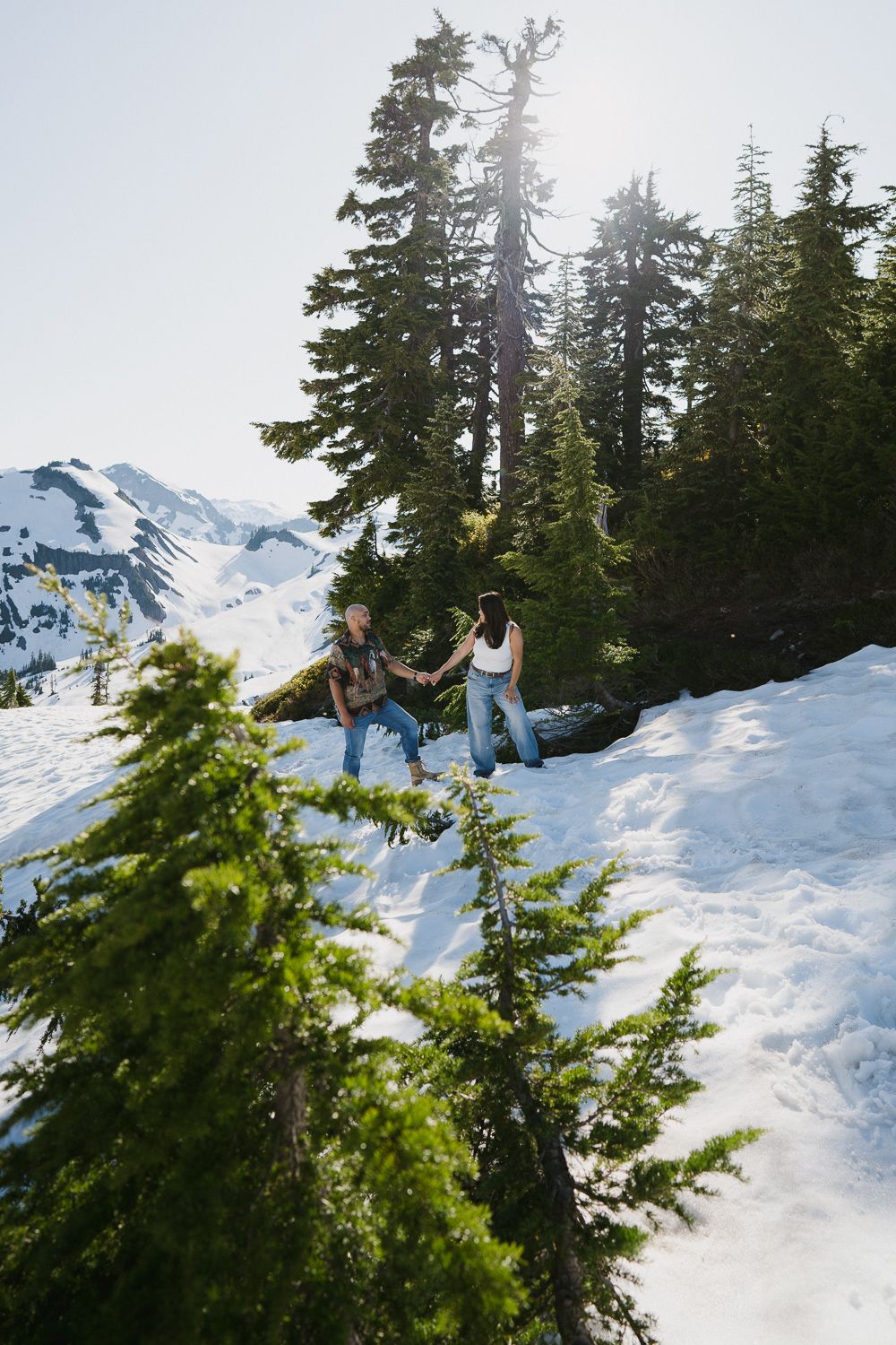 Two people enjoy a winter hike through snowy mountain terrain surrounded by evergreen trees and scenic mountain peaks.