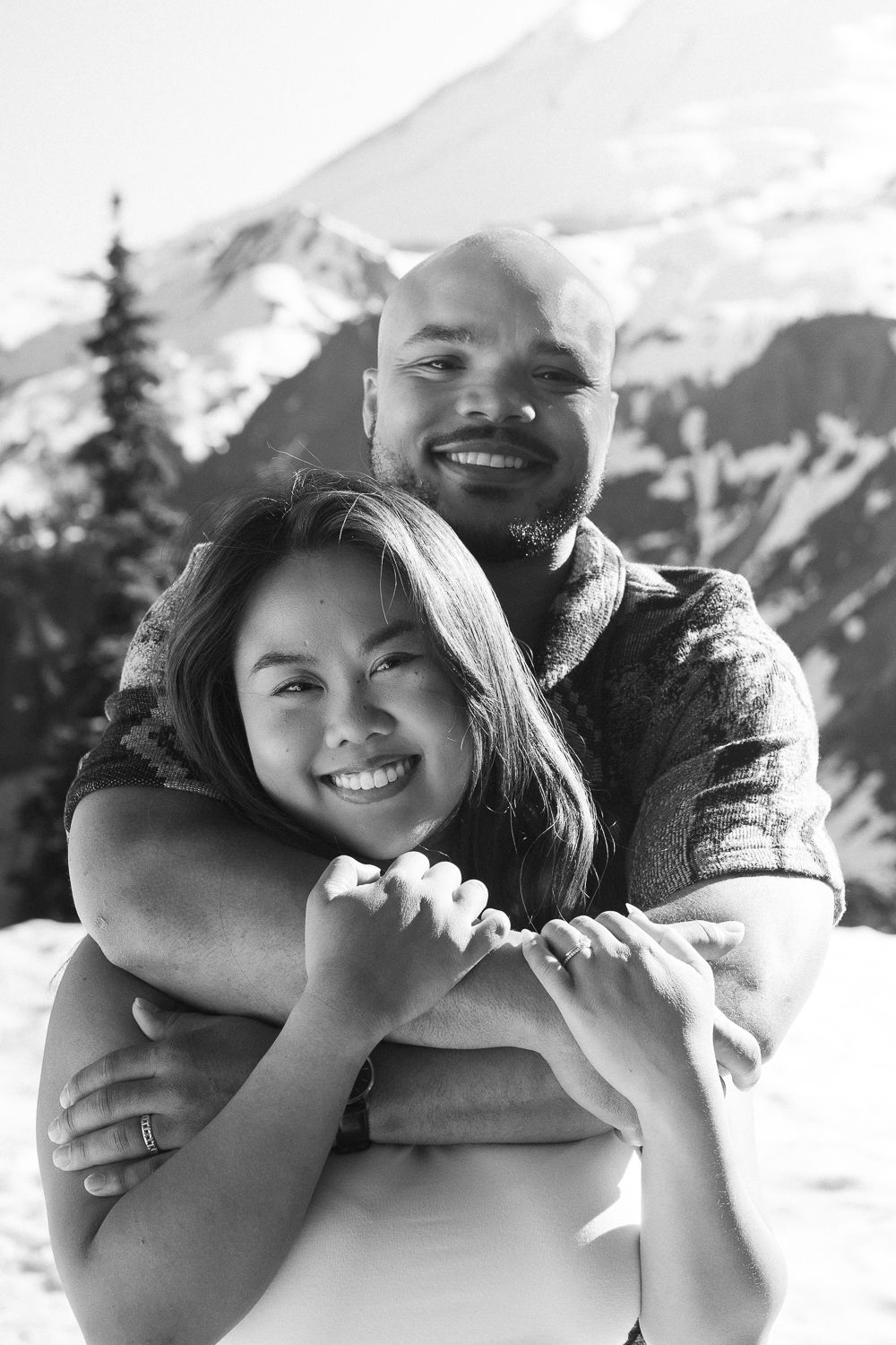 A couple embraces and smiles while posing in front of snowy mountain peaks and evergreen trees.