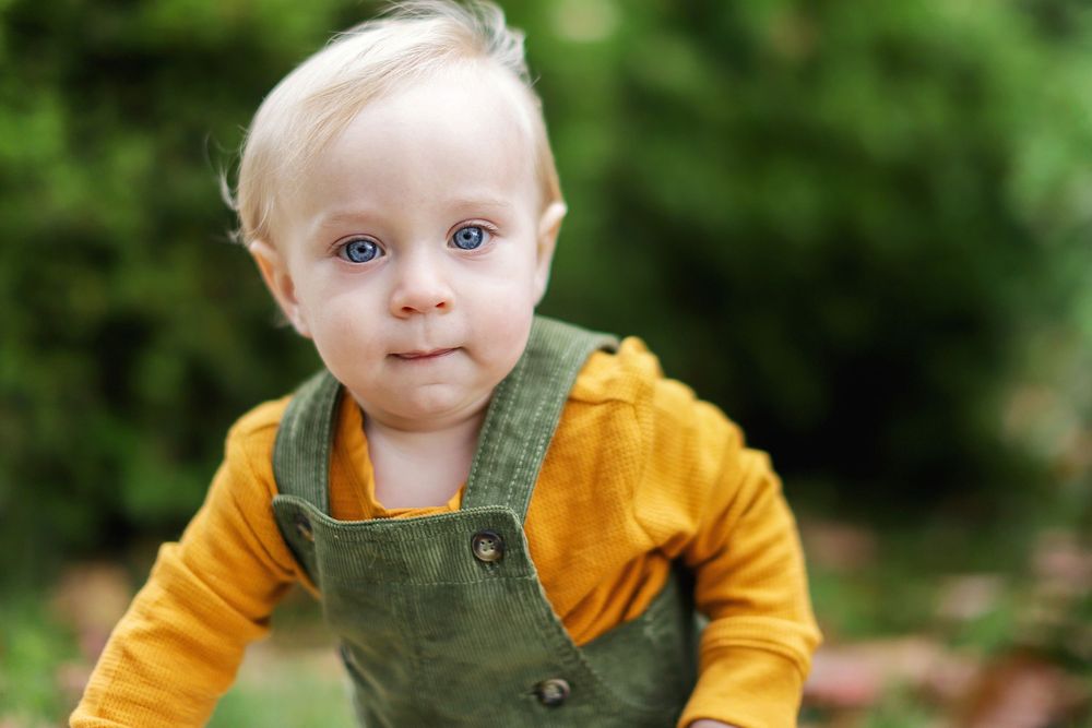 A baby wearing a mustard yellow sweater and olive green overalls sits outdoors surrounded by greenery and natural light.