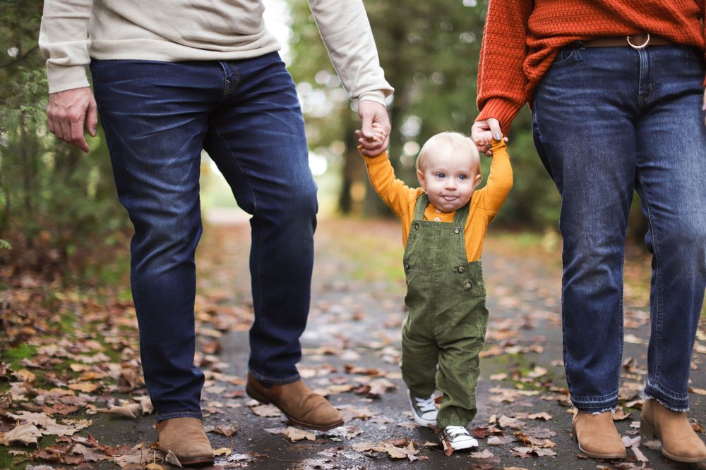 A small child wearing green overalls holds hands with their parents while walking on a leaf-covered path in autumn.