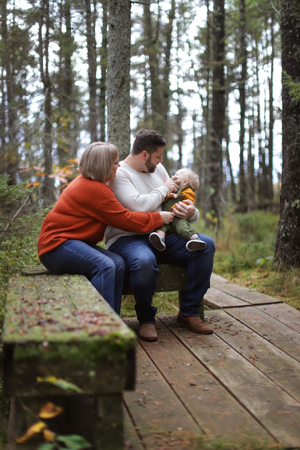 Two people sitting on a wooden walkway in forest share an intimate moment among pine trees and fallen leaves.