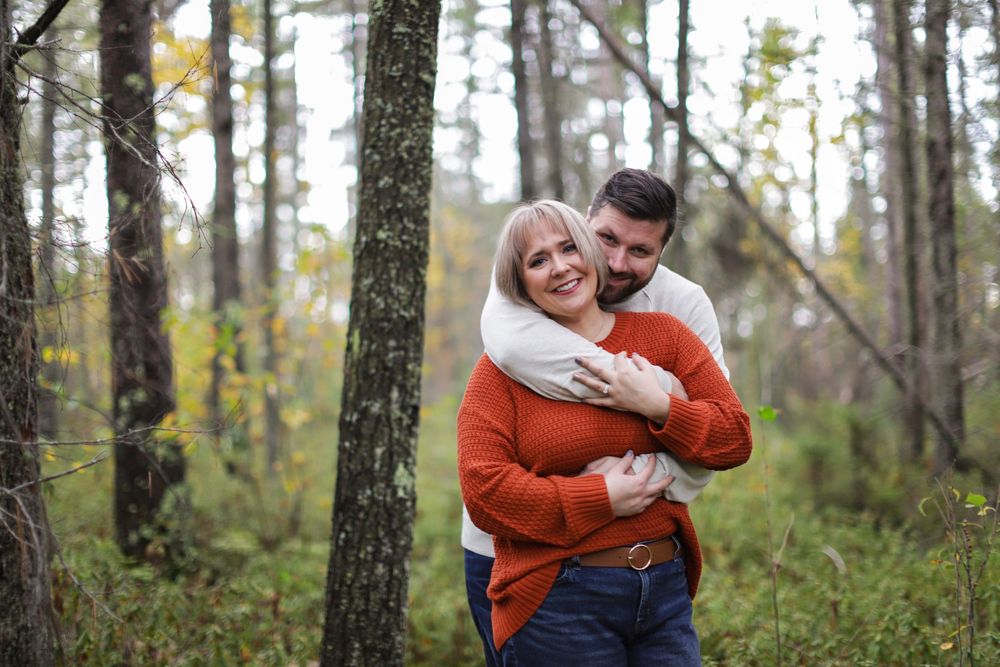 A couple shares an intimate embrace while standing in a wooded forest setting during autumn.