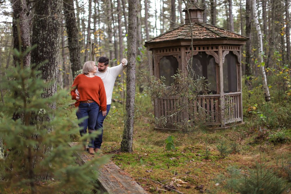 A wooden gazebo stands in a pine forest while two people walk along a narrow dirt path in autumn.