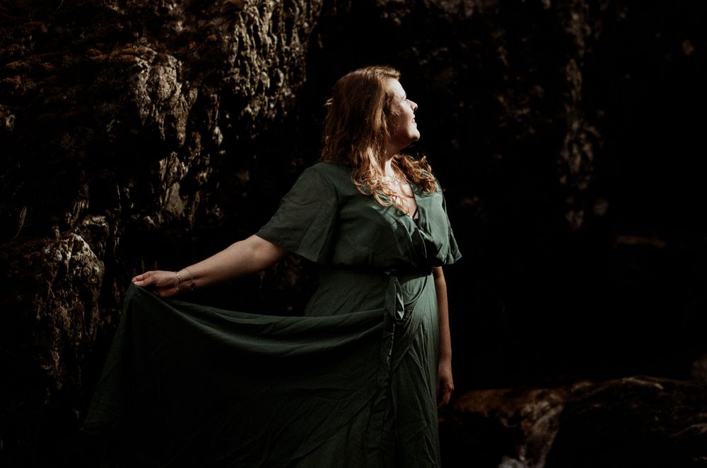 Person in dark green dress poses dramatically in dim lighting while holding flowing fabric against moody backdrop.
