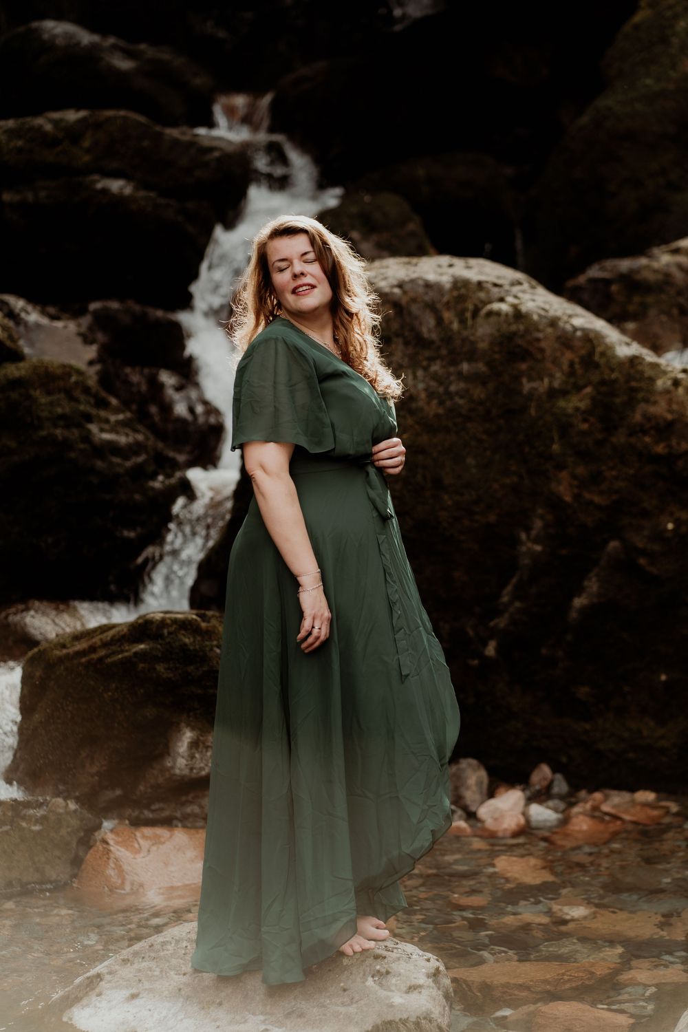 Someone in a flowing dark green dress poses gracefully near a rocky waterfall in natural outdoor setting.