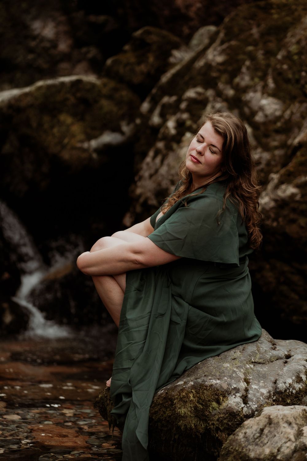 Person in green dress sits peacefully on a rock by a waterfall in a moody forest setting.