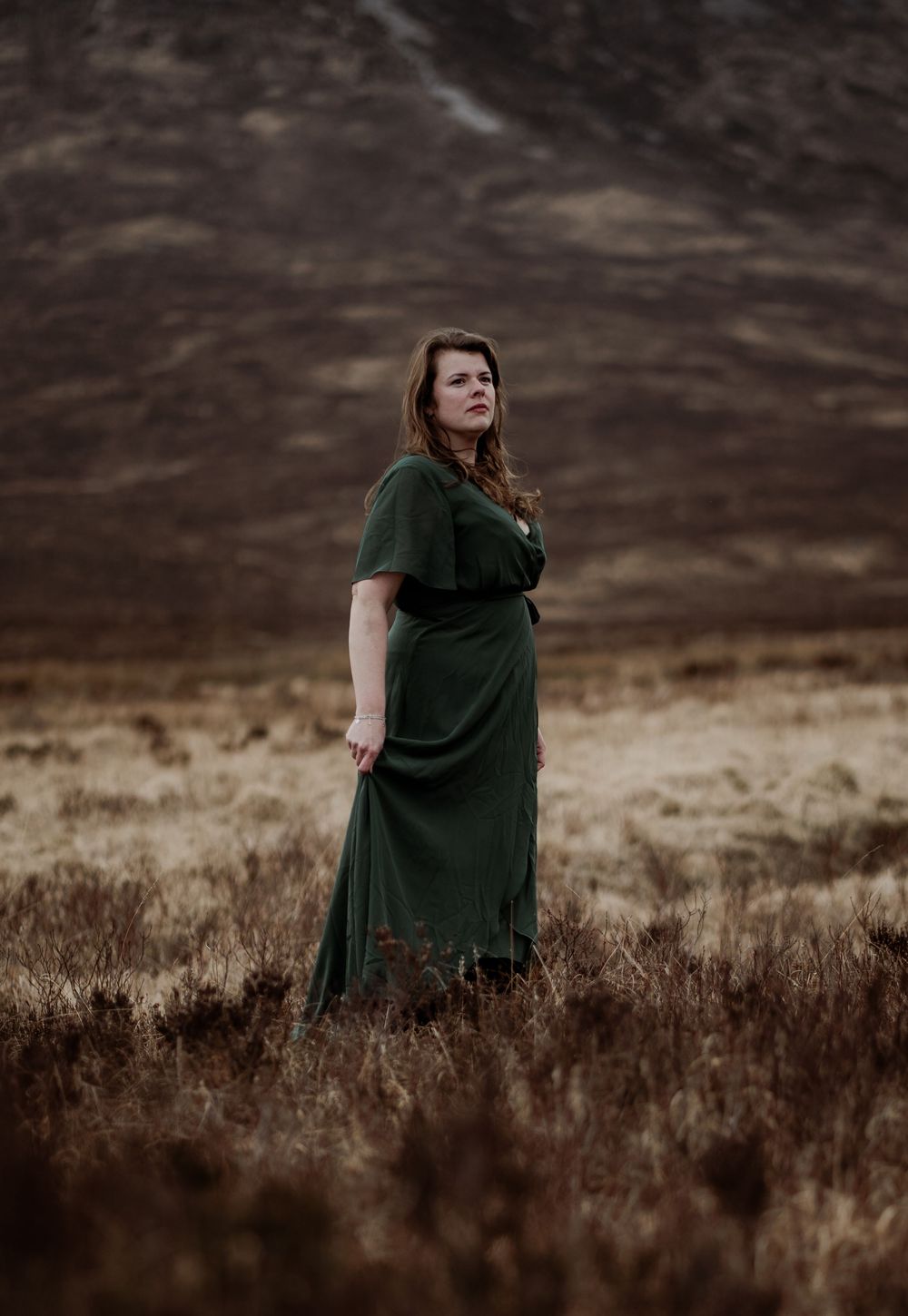 Figure standing in dark green dress against moody mountainous landscape with brown heather in foreground.