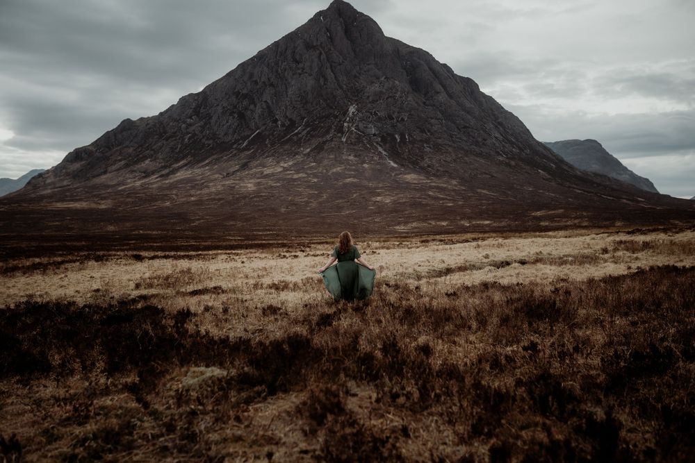 A figure in a green coat stands in the rugged Scottish Highlands beneath the dramatic Buachaille Etive Mor mountain.
