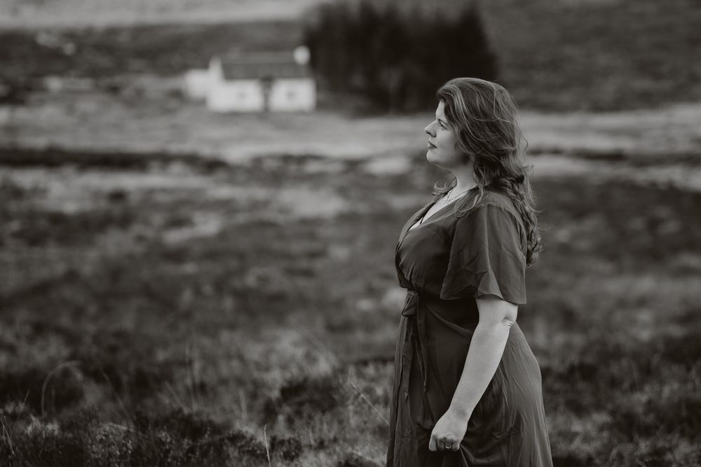 A moody black and white portrait of someone in a dress standing in a highland landscape with a white cottage in the distance.