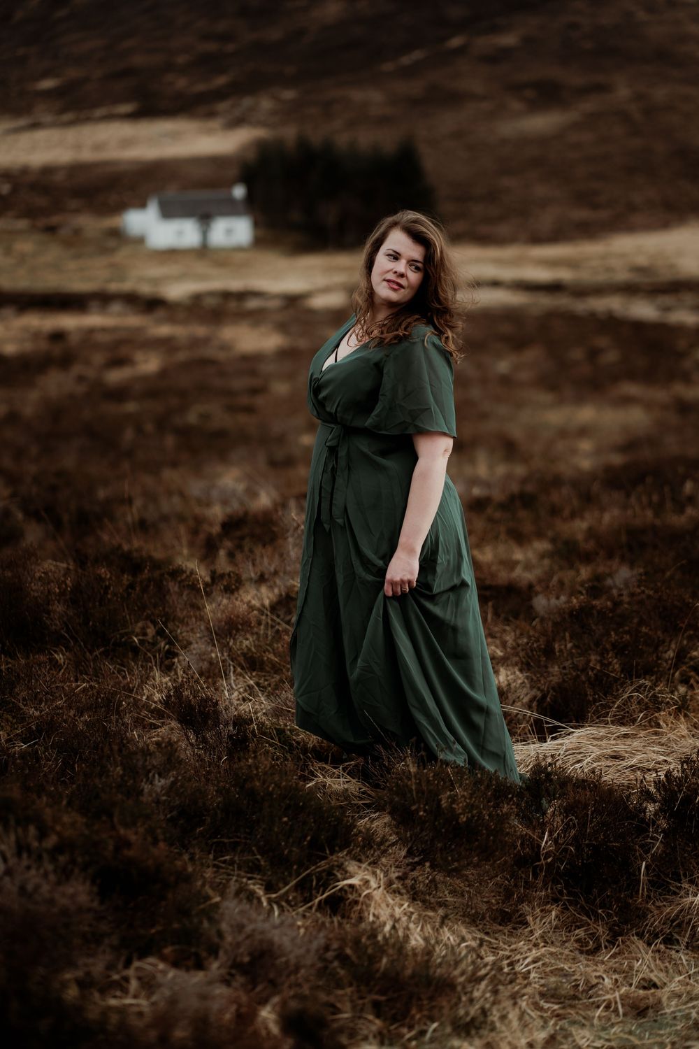 Person in emerald green dress sits gracefully on moorland terrain with white cottage visible in distant background.